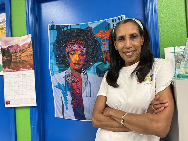 African American woman in a white t-shirt standing by a work of fabric art showing a Black female doctor