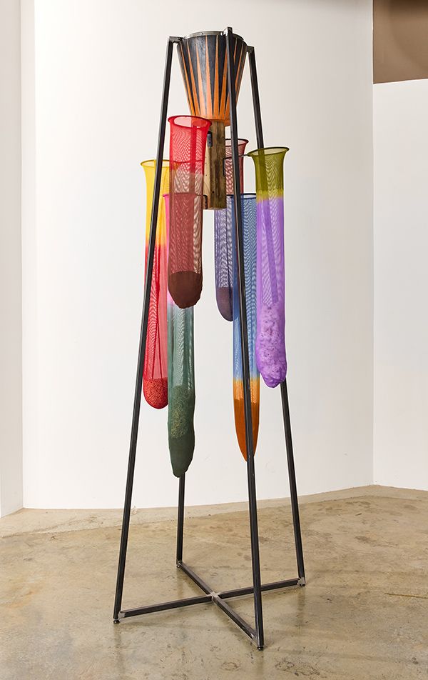 Tall, multicolored sculpture made from a steel frame, a wooden drum-like object, and multiple nylon nets holding agricultural products such as tobacco and rice